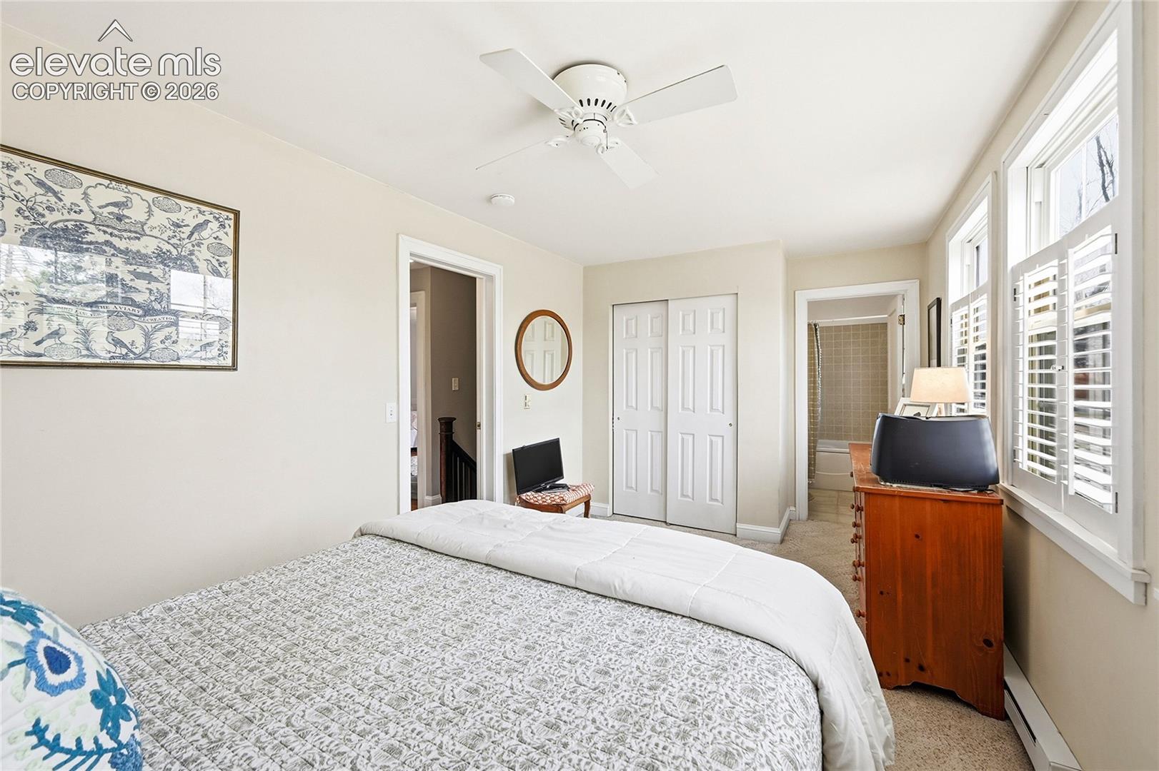 Neutral-toned interior featuring dual windows with adjustable shutters, a ceiling fan, and a double-door closet