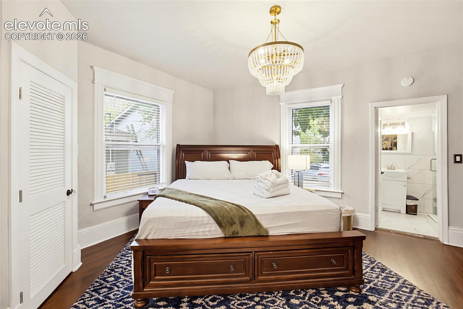 Hardwood flooring, white louvered closet door, decorative ceiling light fixture, white window blinds, and an attached three-quarter bathroom with a walk-in shower