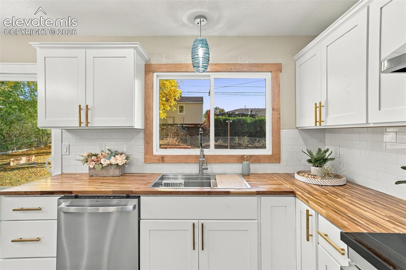 White shaker cabinetry with brass hardware, butcher block countertops, subway tile backsplash, stainless steel dishwasher, and sliding window with wood trim