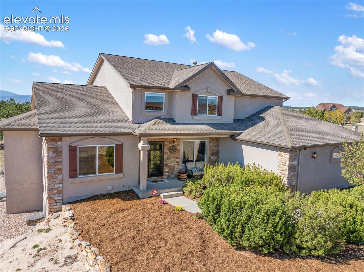 Stucco exterior featuring stacked stone accents, shingle roofing, decorative shutter hardware, and a covered front entry with a column