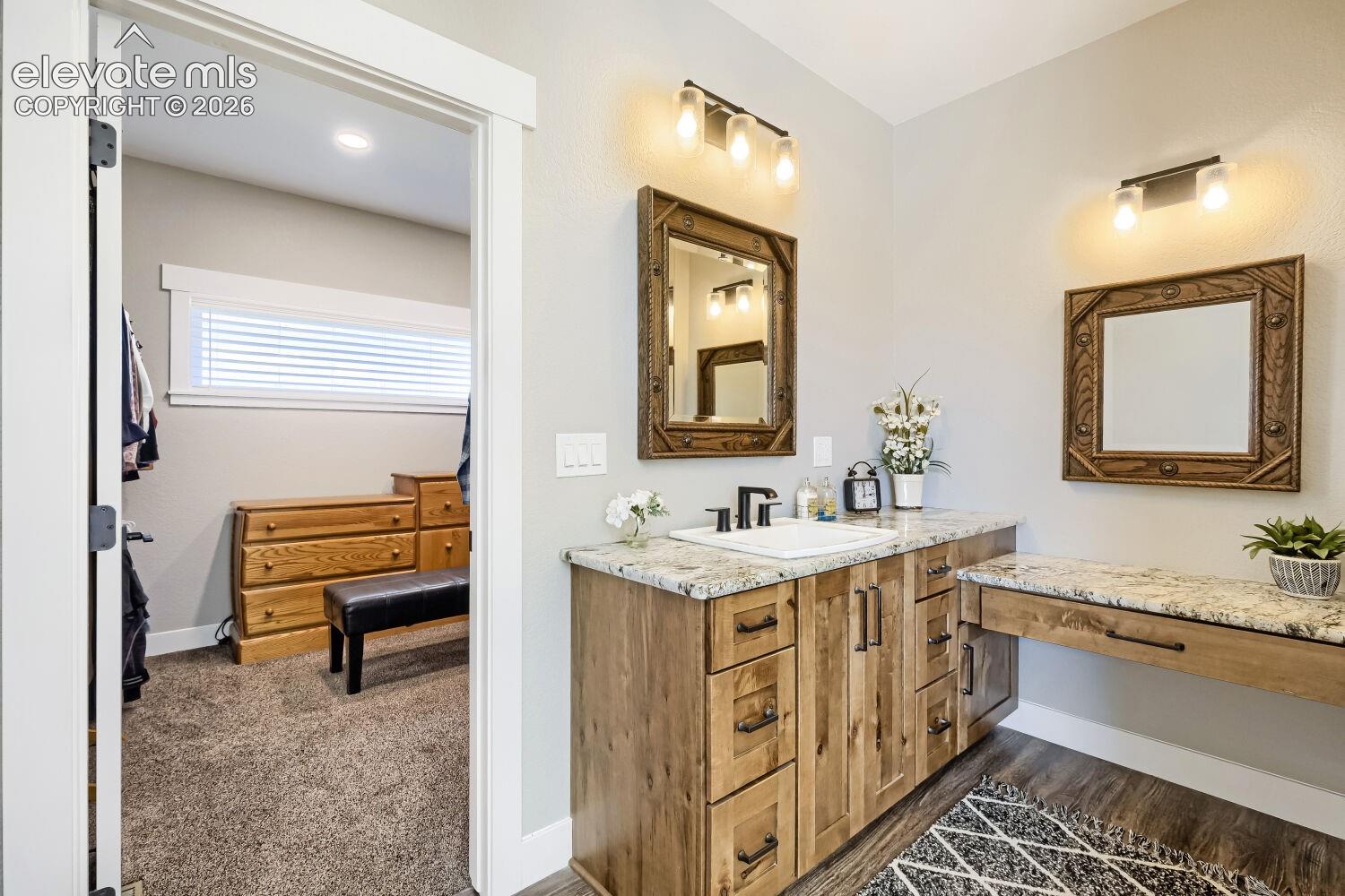 Dual vanity light fixtures, wood-finish flooring, light-toned cabinetry, granite-style countertop, and a built-in makeup vanity