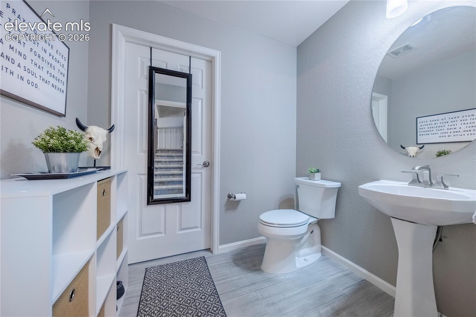 Pedestal sink with polished chrome faucet, large circular wall mirror, wood-finish tile flooring, and six-panel interior door