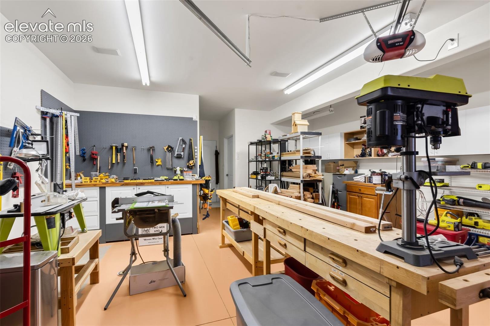 Garage workspace featuring wall-mounted pegboard organization, extended wood-top workbench, white cabinetry, and overhead fluorescent lighting
