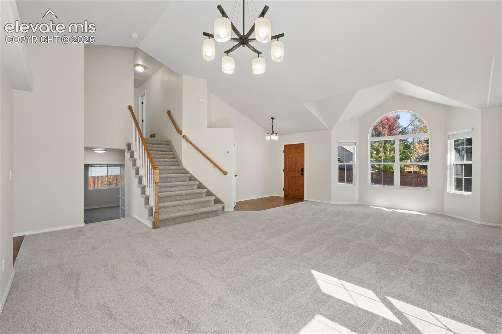 Vaulted ceilings, grand chandelier, arched window, staircase with wooden handrail, and neutral carpeting