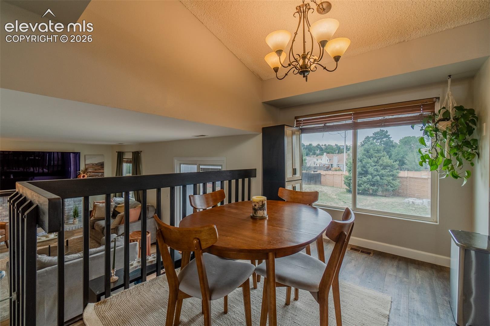 Vaulted ceiling, wood-finish flooring, large window with blinds, railing overlooking lower level, and an overhead chandelier