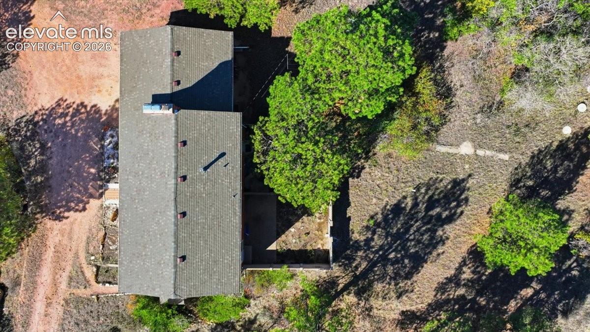 Aerial view of property featuring a shingled roofline, chimney stack, paved driveway, and mature tree coverage on a natural landscape