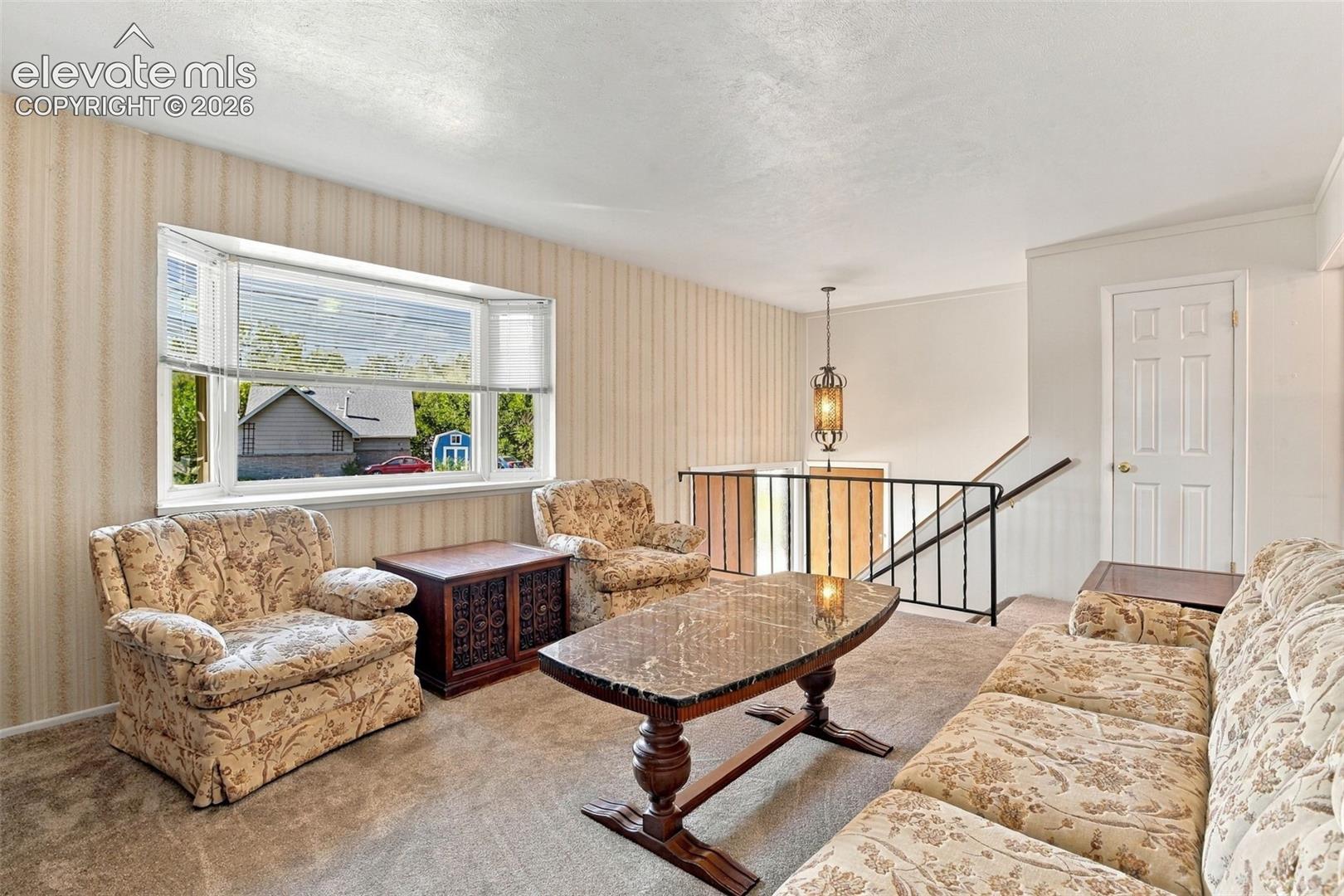 Living area featuring a large bay window, wall-to-wall carpeting, metal stair railing, and a pendant light fixture