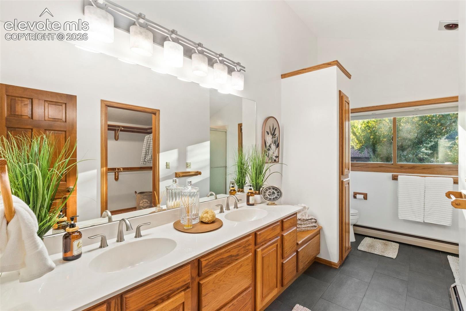 Dual-sink vanity featuring light-toned solid surface countertops, natural wood cabinetry, polished chrome fixtures, and a large-format vanity mirror with integrated lighting