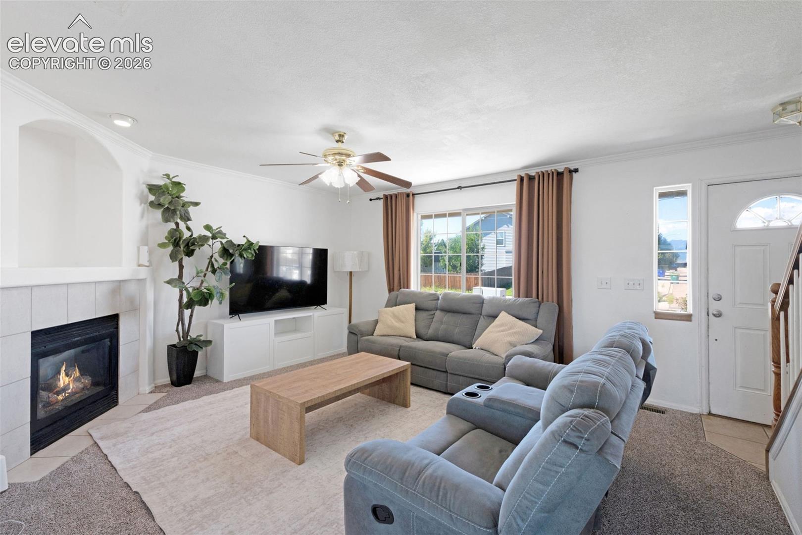 Living area featuring a fireplace with a tiled surround, a large window with brown drapes, and a ceiling fan with light fixture