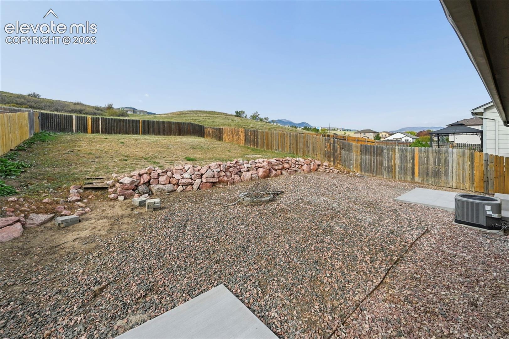 Fenced backyard featuring natural stone retaining walls, gravel landscaping, concrete patio sections, and mountain backdrop