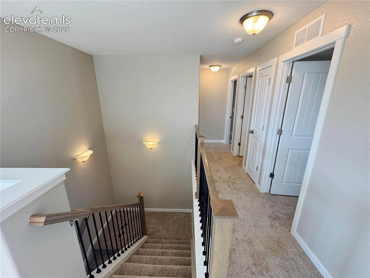 Hallway featuring carpeted flooring, two-panel interior doors, wall-mounted sconce lighting, and a staircase with a wooden handrail and metal balusters