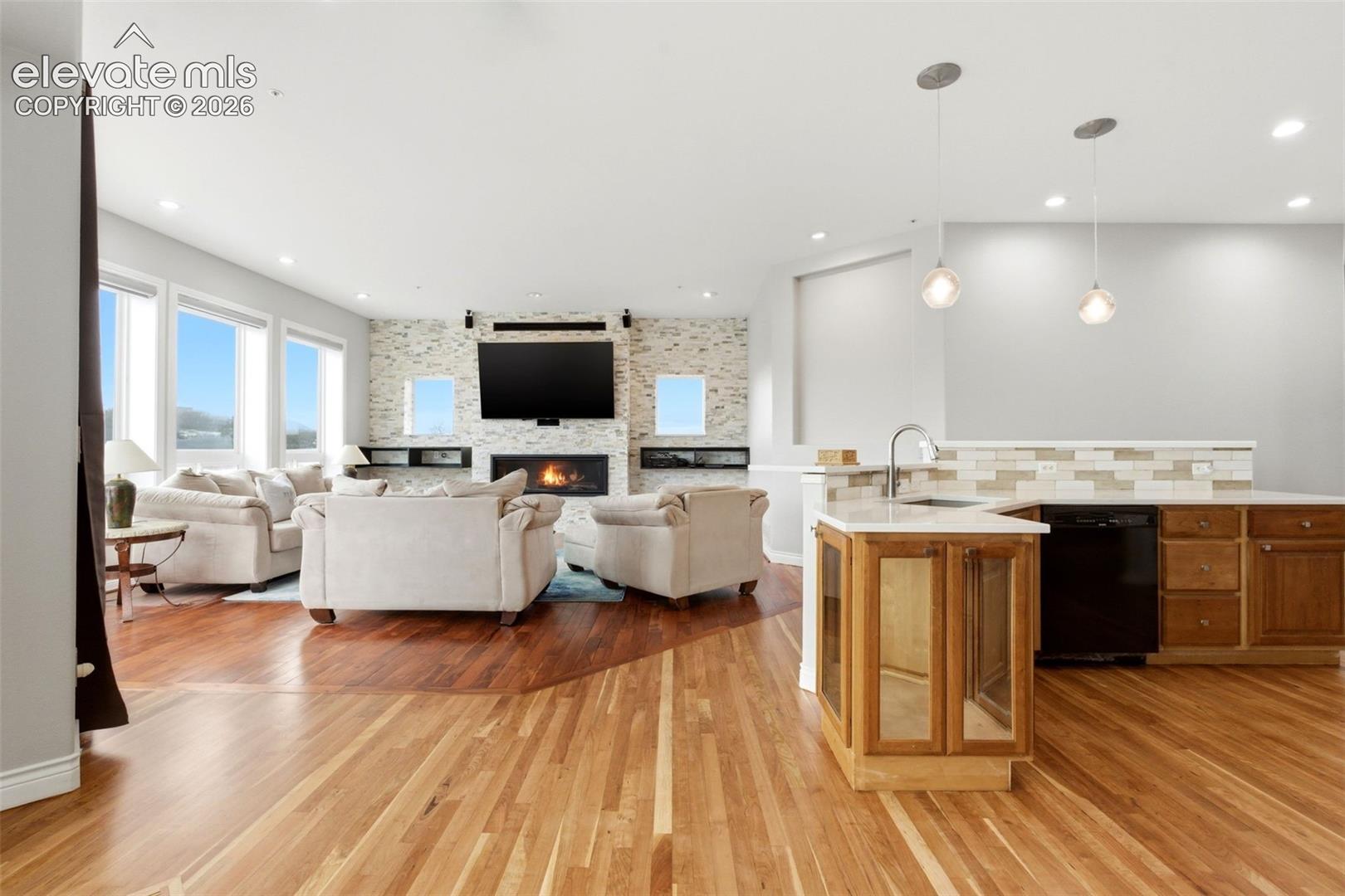 Hardwood flooring throughout, a stacked stone fireplace, recessed lighting, multiple large windows, and a kitchen island with a sink and pendant lighting