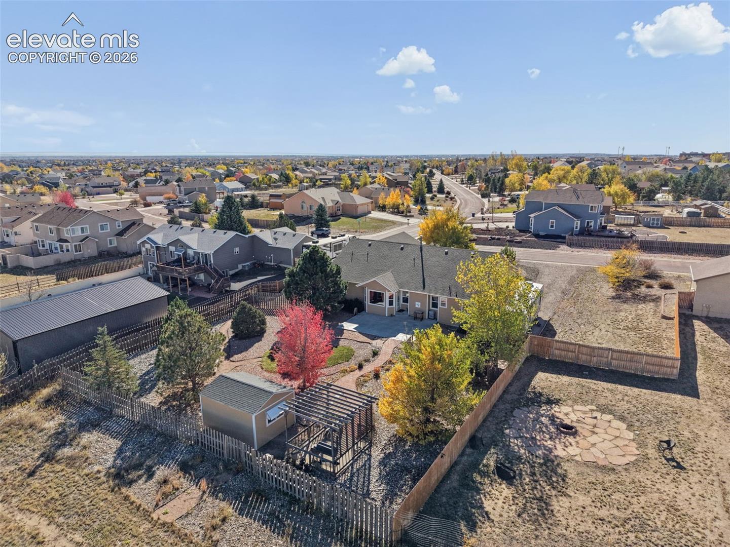 Aerial perspective of a property featuring a fenced backyard, a detached storage shed, a pergola, a stone-paved fire pit area, and mature trees
