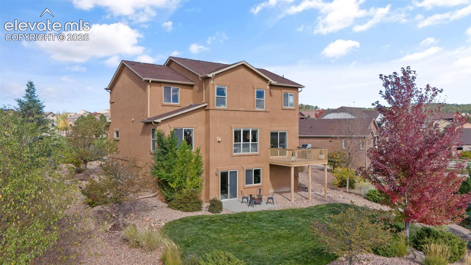 Multi-story exterior featuring a stucco facade, elevated wood-tone deck, paved lower-level patio, and landscape design incorporating mature trees and rock ground cover
