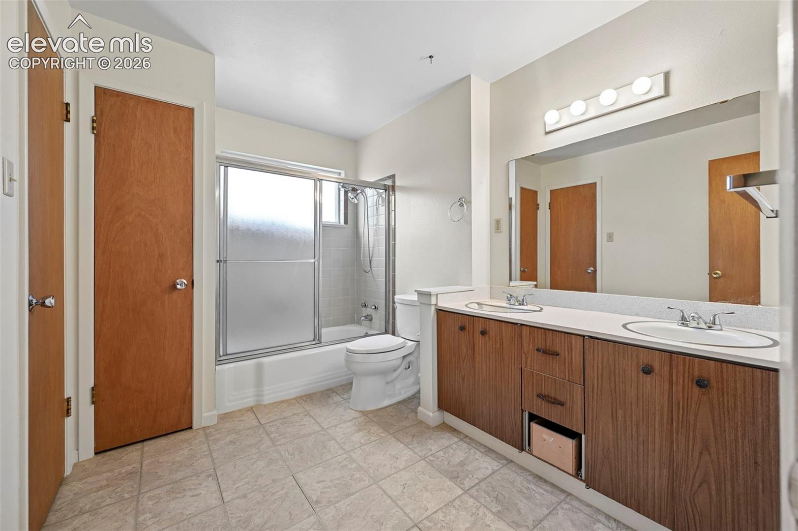 Primary bathroom with double-sinks, with wood-finish cabinetry, a full-width mirror, tile flooring, and a tub-shower combination with sliding glass doors