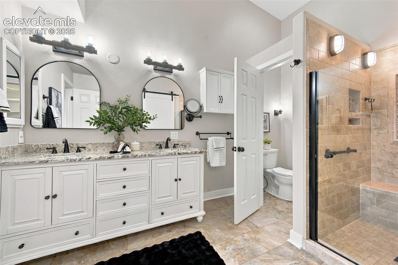 Bathroom featuring a dual-sink vanity with stone countertop, arched mirrors, white cabinetry, and a walk-in shower with tiled walls and a built-in bench