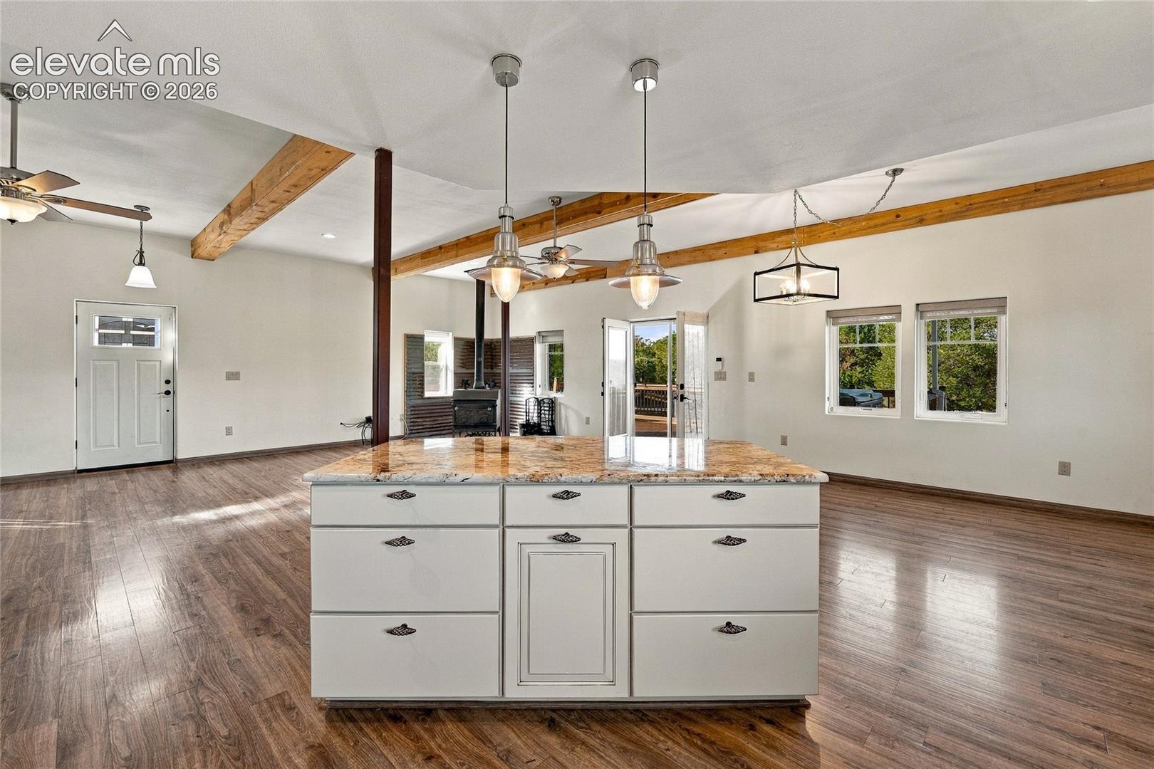 Center island with stone countertop and white cabinetry, wood-finish flooring, exposed structural wood ceiling beams, pendant lighting, and metal-cased wood stove