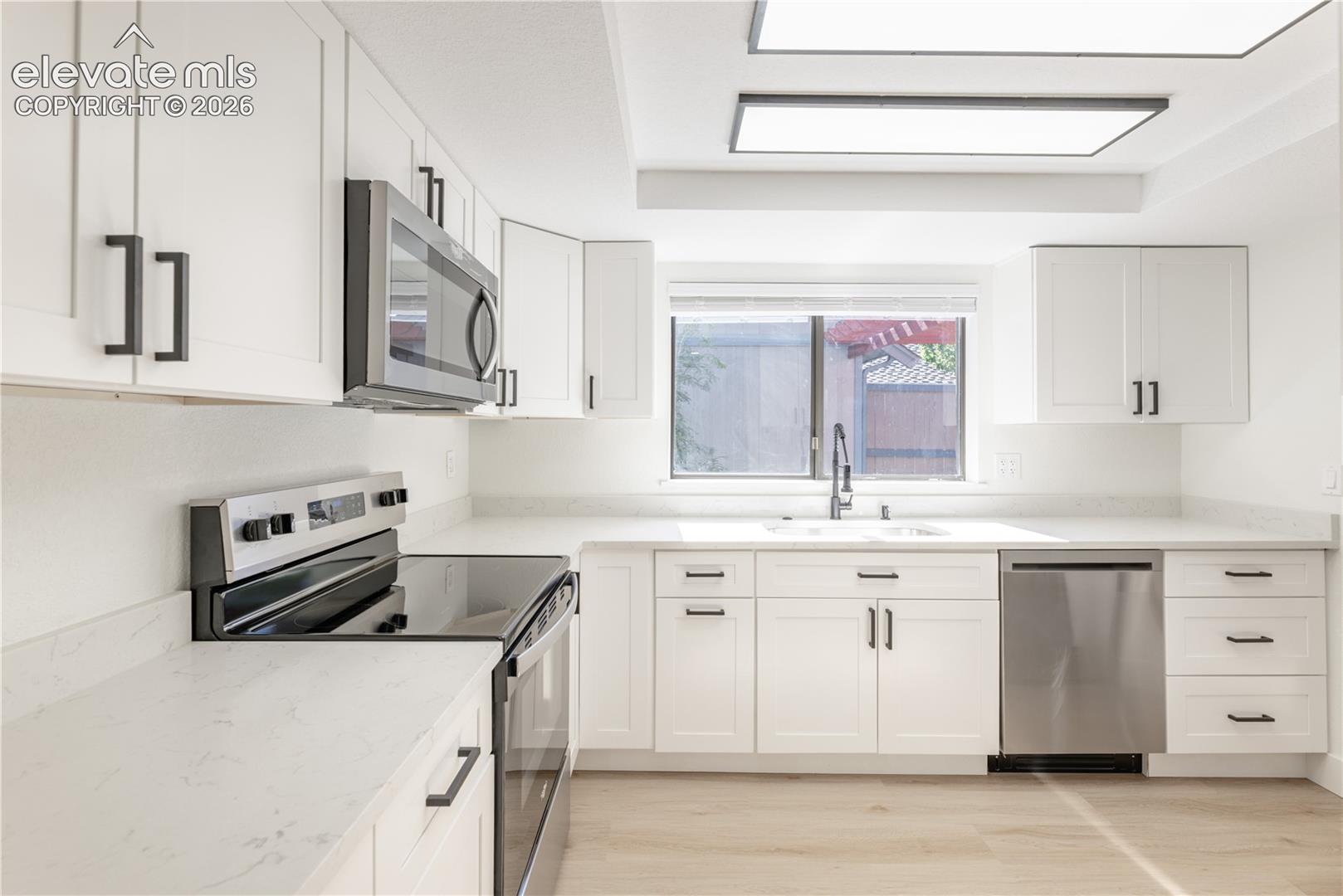 Kitchen featuring white shaker-style cabinetry, quartz countertops, stainless steel appliances, dual-basin undermount sink with a gooseneck faucet, and wood-finish flooring