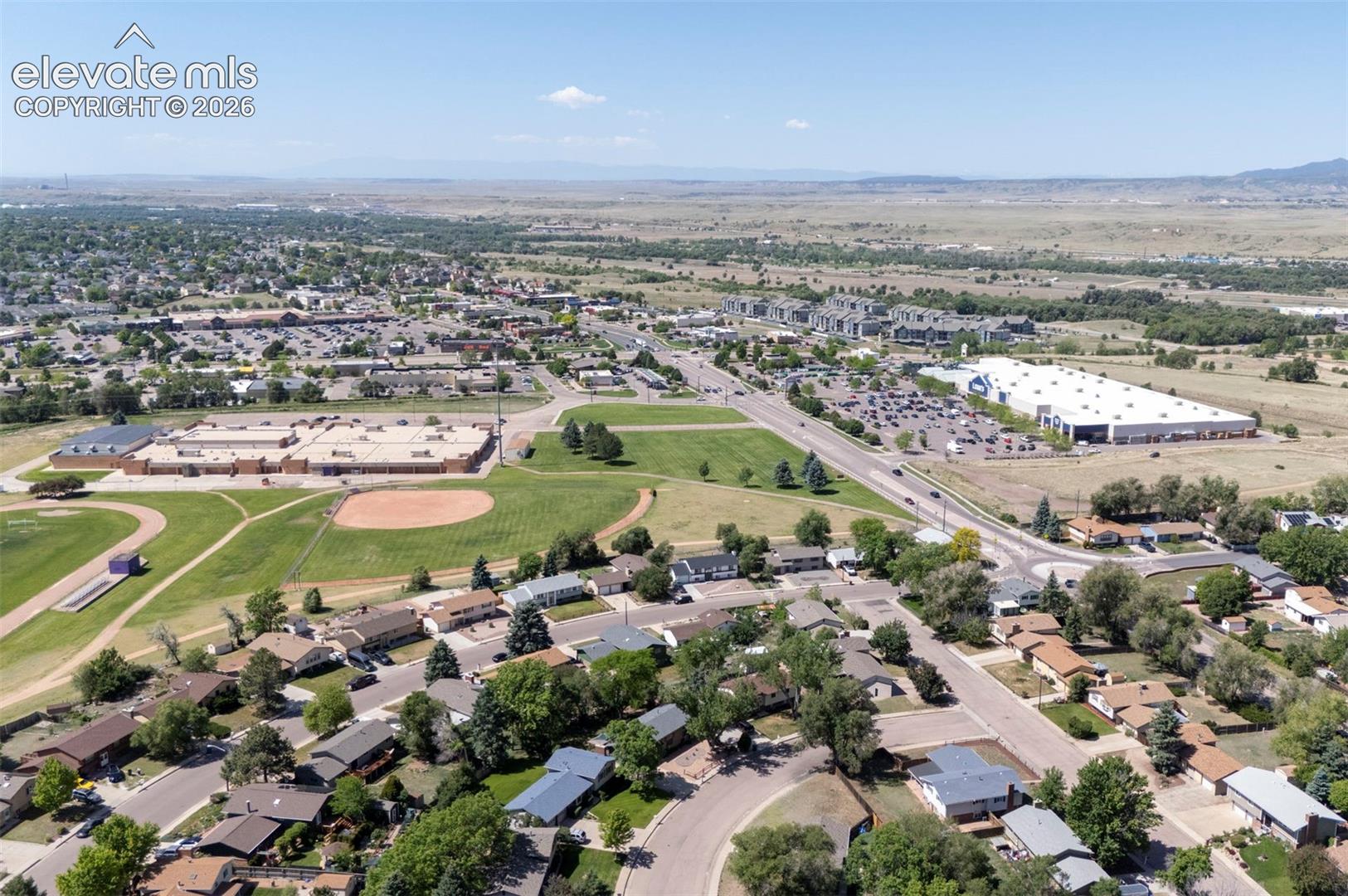 Aerial perspective showcasing a residential suburban landscape featuring paved roadways, mature deciduous trees, expansive grassy fields, and proximity to regional infrastructure and retail centers