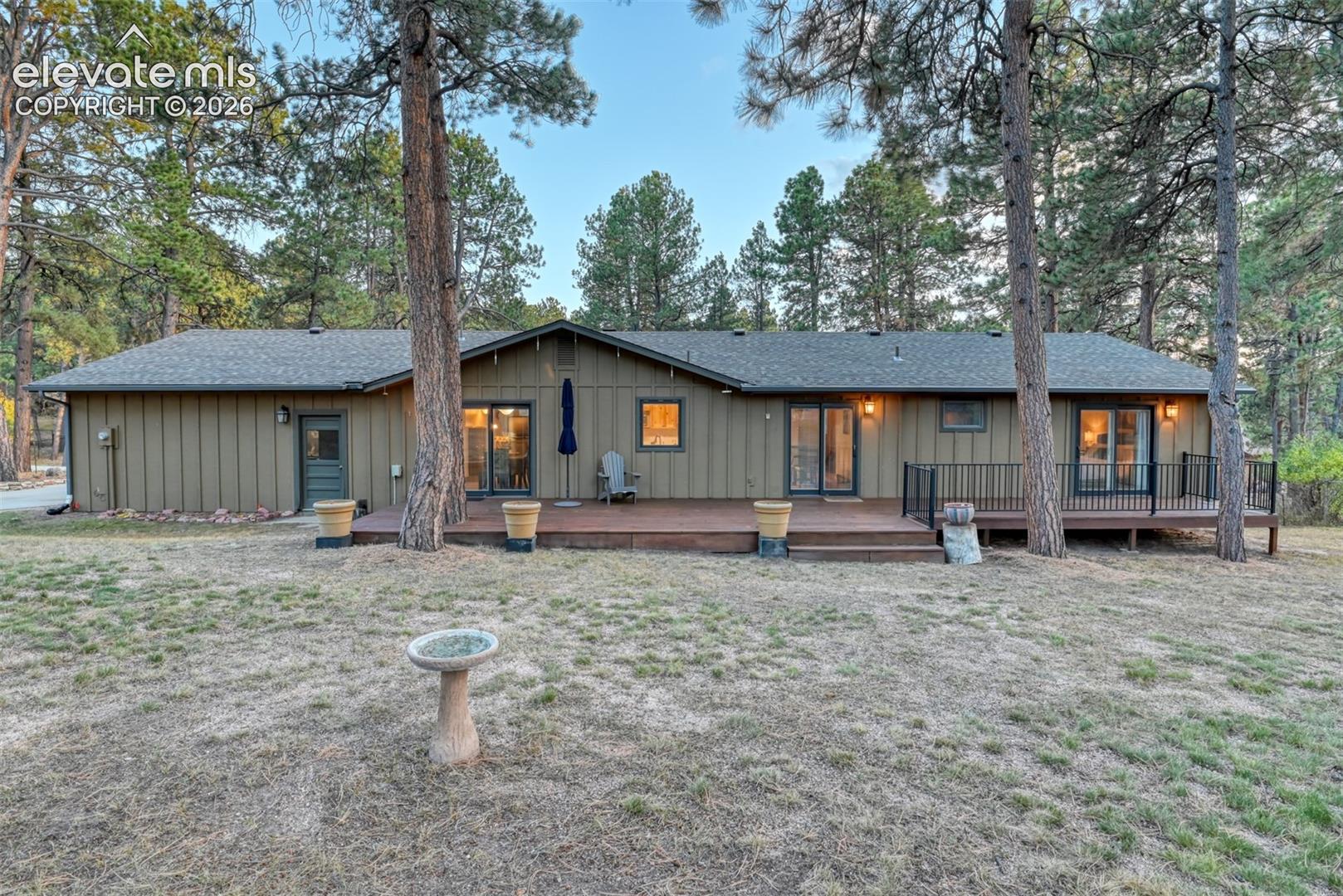 Back exterior with a sprawling rear trex deck with black metal railing and mature pine trees. Slider doors can be seen in the dining room, kitchen nook, and master bedroom.