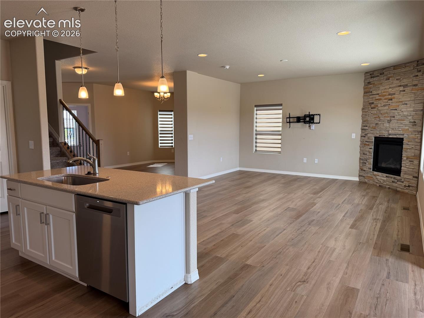 Open-concept living area featuring wood-finish flooring, a kitchen island with a built-in sink and stainless steel dishwasher, a stacked stone fireplace, and recessed ceiling lighting
