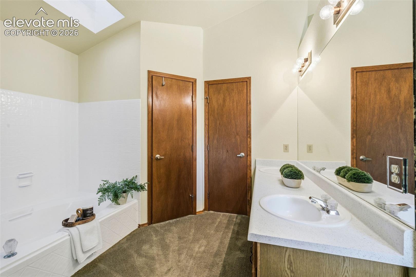 Bathroom featuring a dual-sink vanity with a solid-surface countertop, a soaking tub with integrated surround, a vaulted ceiling with a skylight, and wall-mounted vanity lighting