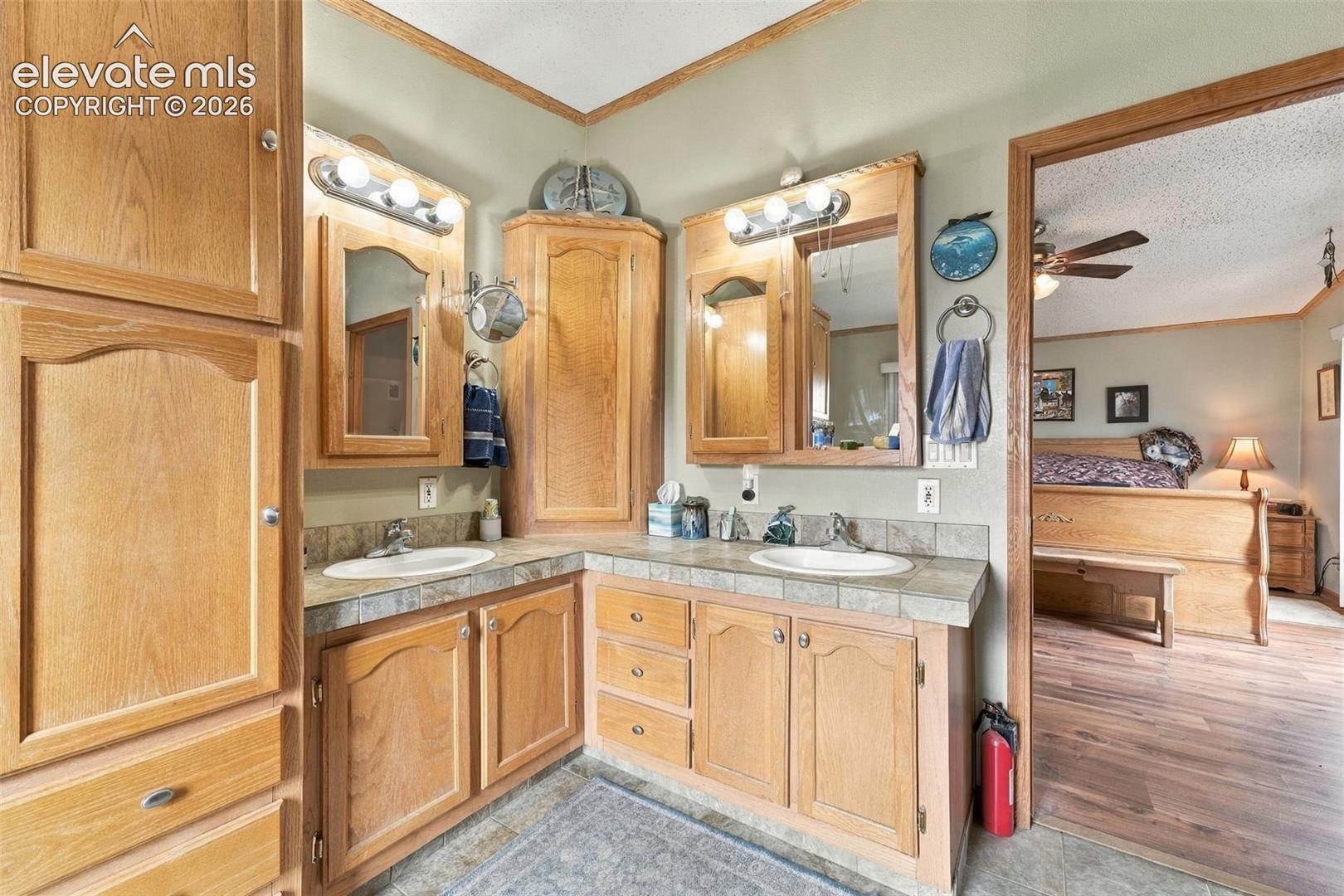 Dual-sink vanity with light wood cabinetry, tile countertops, corner storage tower, and dual-bulb lighting fixtures