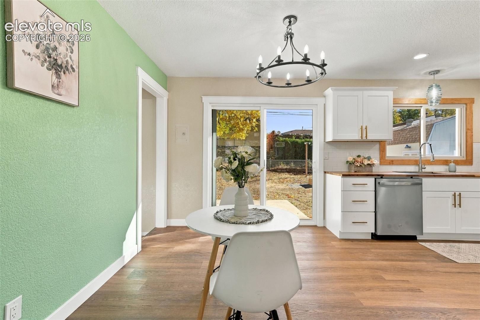 Kitchen and dining area featuring white shaker-style cabinetry, stainless steel dishwasher, wood-finish flooring, wrought-iron chandelier, and sliding glass door