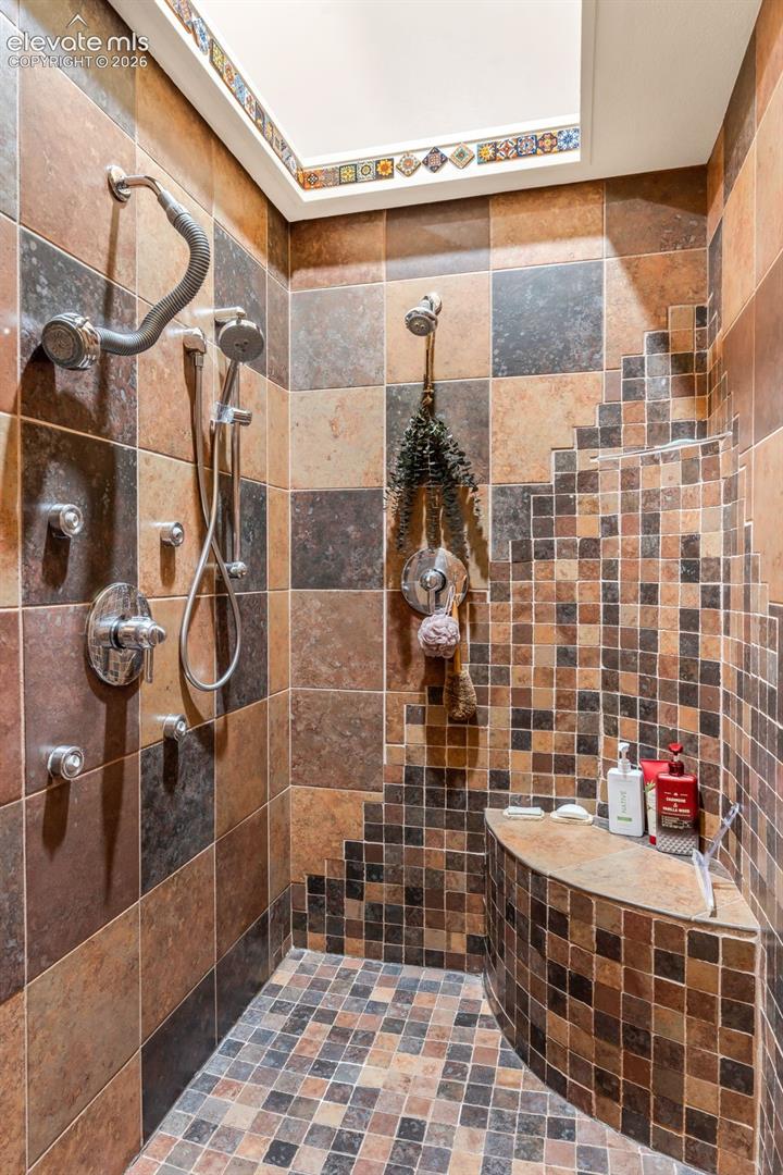 Walk-in shower featuring multi-tone square tile surround, built-in curved tile bench, dual shower heads, integrated body sprays, and a decorative tile-bordered skylight