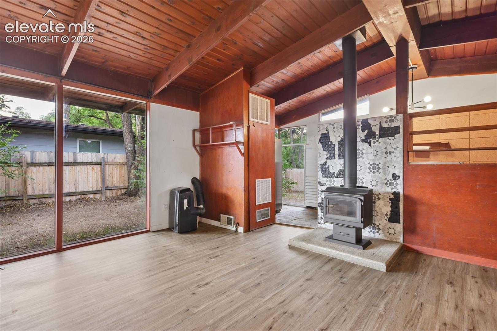 Vaulted wood-paneled ceilings with exposed beams, full-height windows, wood-finish flooring, wood-burning stove with a tile surround and hearth, and built-in shelving