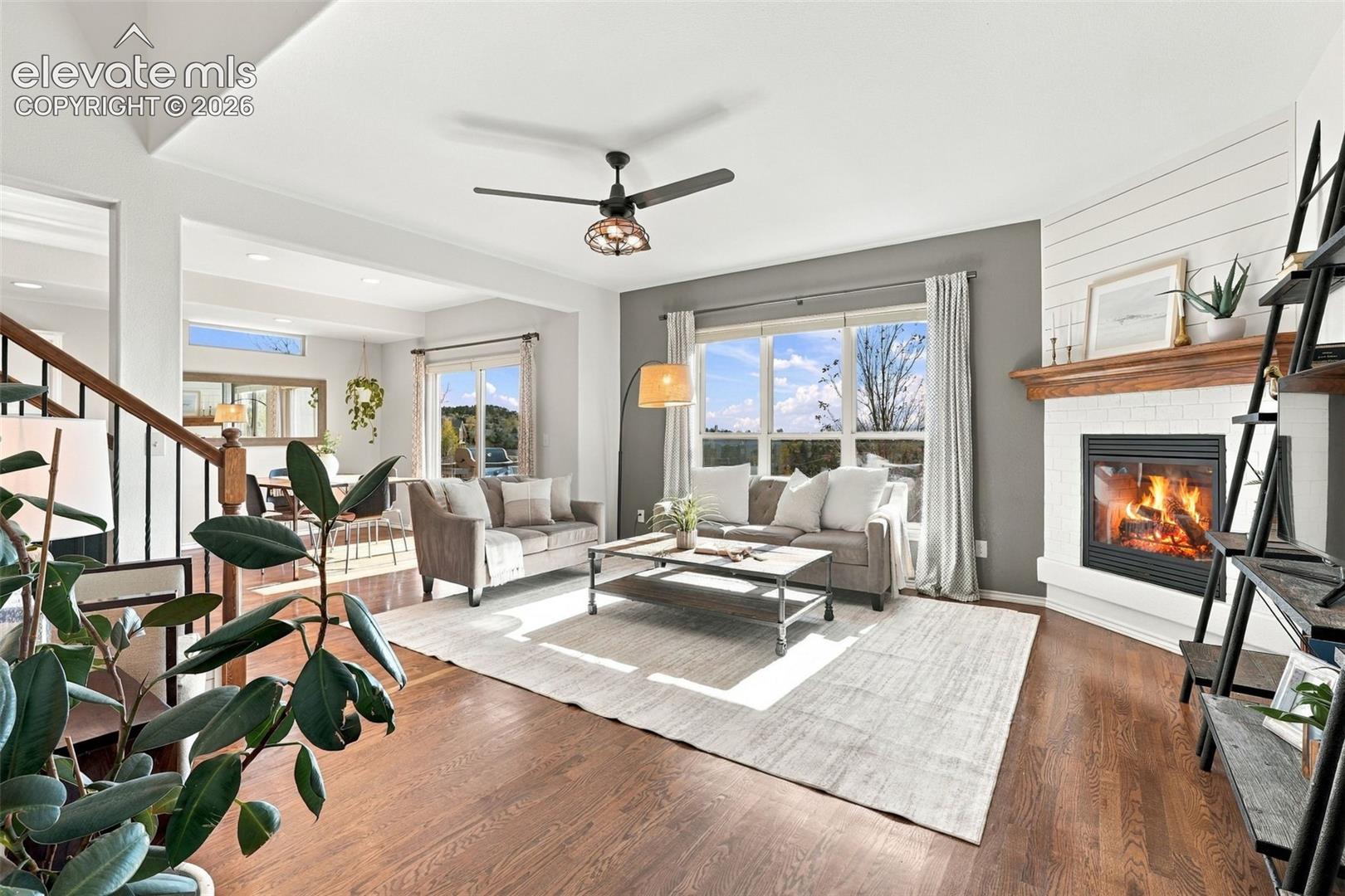 Living area featuring wood-finish flooring, a wood-burning fireplace with a white brick surround, a rustic wood mantel, white shiplap wall accents, and a ceiling fan