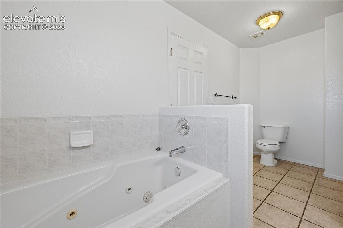 Soaking tub with chrome fixtures, tiled surround, white paneled door, ceramic tile flooring, and a standard toilet