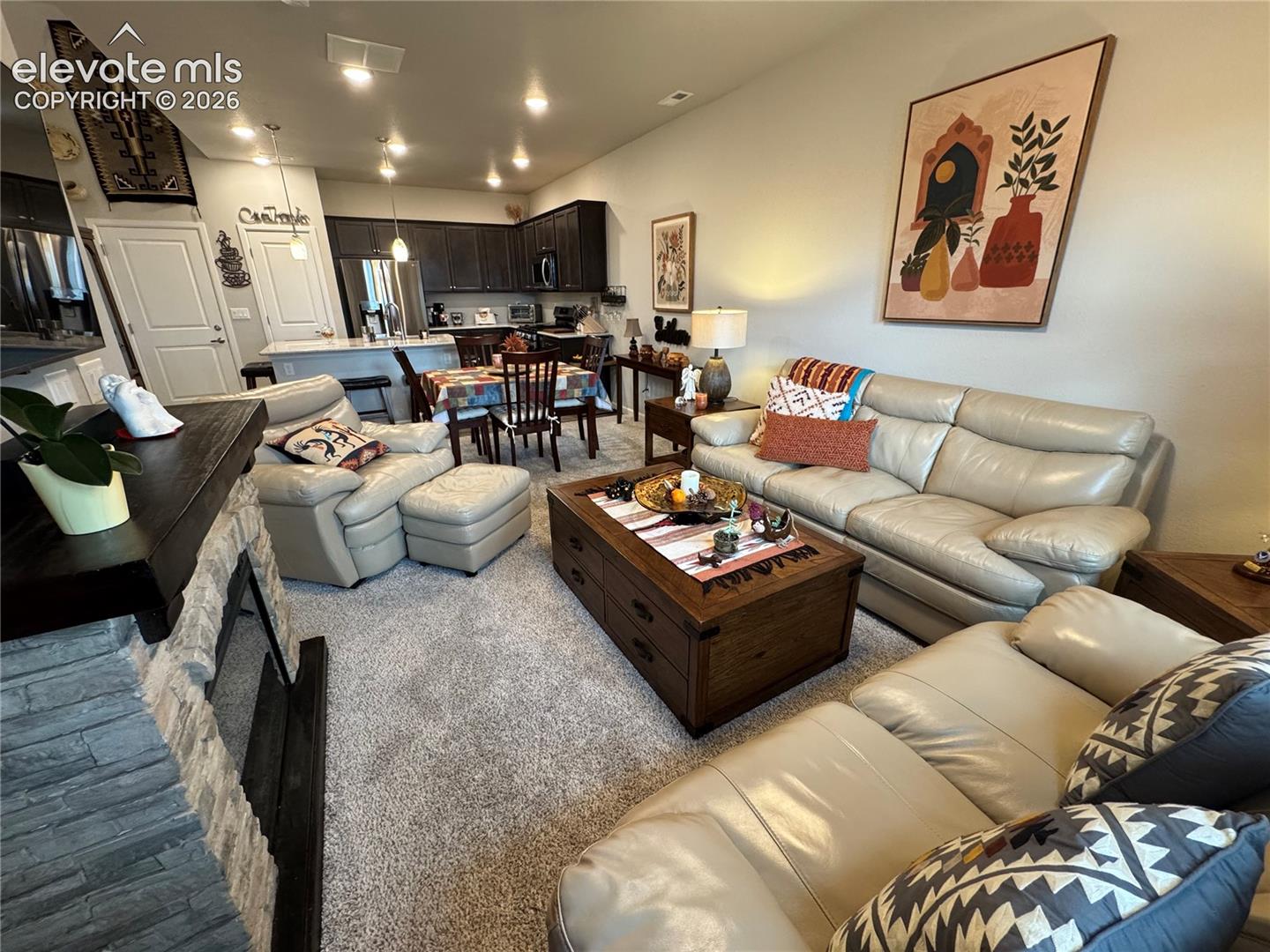 Open-concept area featuring a floor-to-ceiling stone-veneer fireplace, kitchen with dark-toned cabinetry, breakfast bar, and recessed lighting