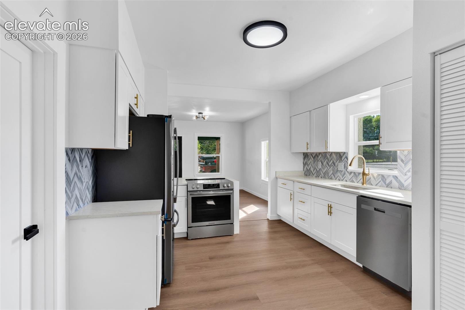 Kitchen featuring white Shaker-style cabinetry, stainless steel appliances, herringbone-patterned tile backsplash, gold-tone hardware, and wood-finish flooring
