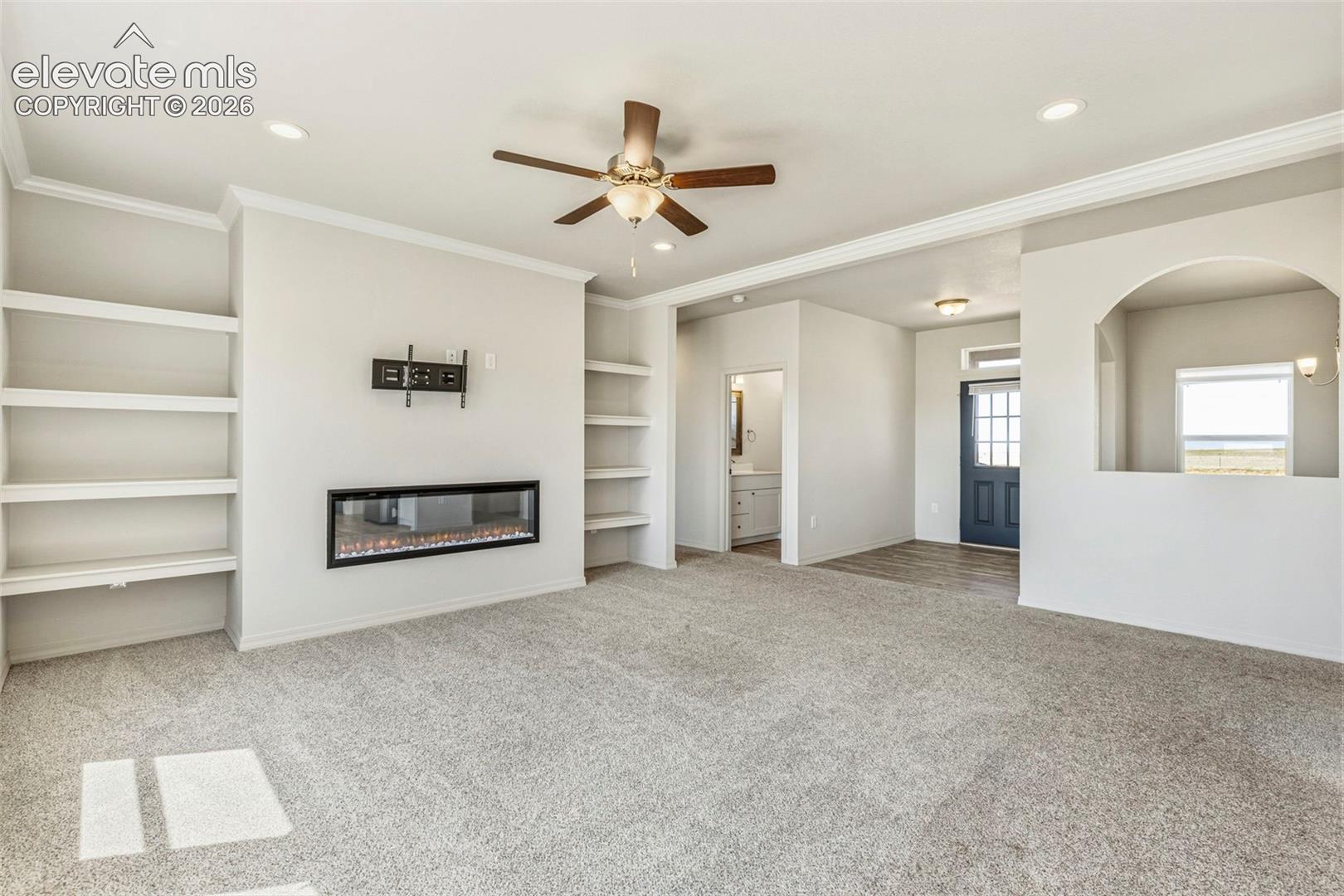 Spacious living area featuring an electric fireplace with a wall-mounted television bracket, built-in shelving, recessed lighting, and a ceiling fan