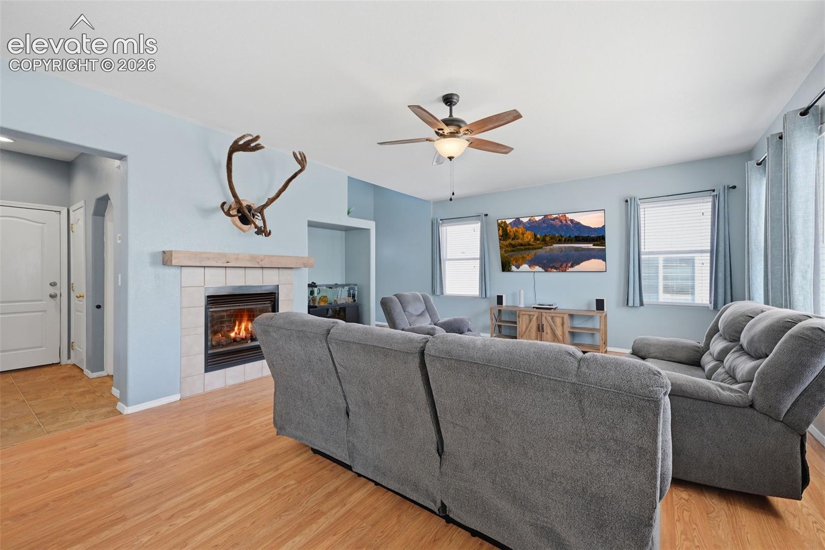 Living space featuring wood-finish flooring, a tiled fireplace with a wood mantle, and a ceiling fan with integrated lighting