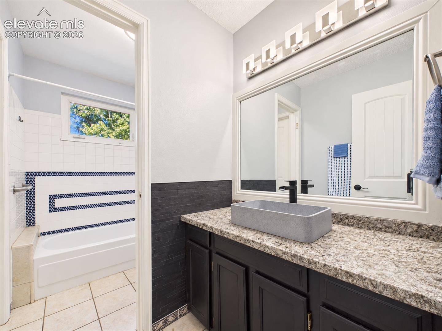 Bathroom vanity pairs dark cabinetry with a stone-look countertop, rectangular vessel sink, oversized mirror, contemporary lighting, and contrasting wall accents.