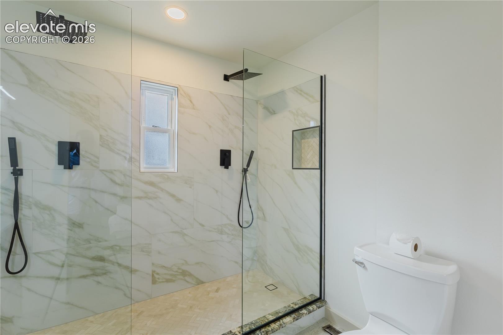 Spacious walk-in shower with floor-to-ceiling glass enclosure, large format marble-look tile, dual matte black showerheads, and a recessed shower niche