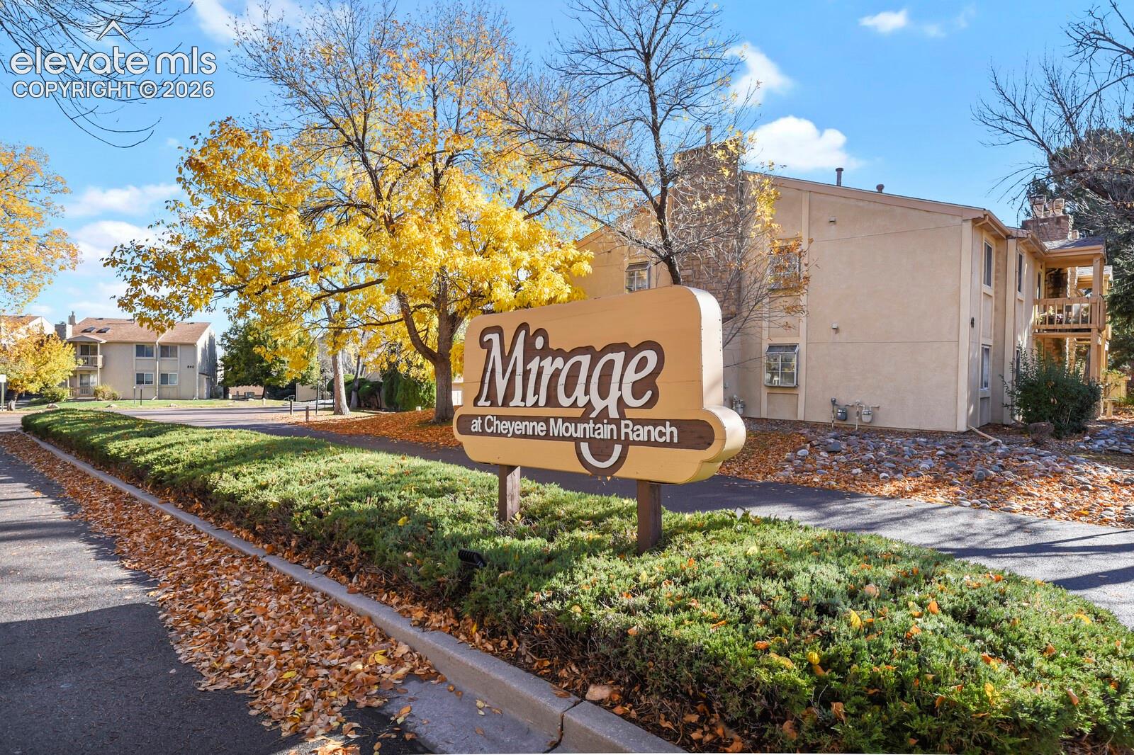 Entrance to the Area showing community signage featuring structured landscaping, mature trees, and multi-story building exteriors with stucco siding and private balconies