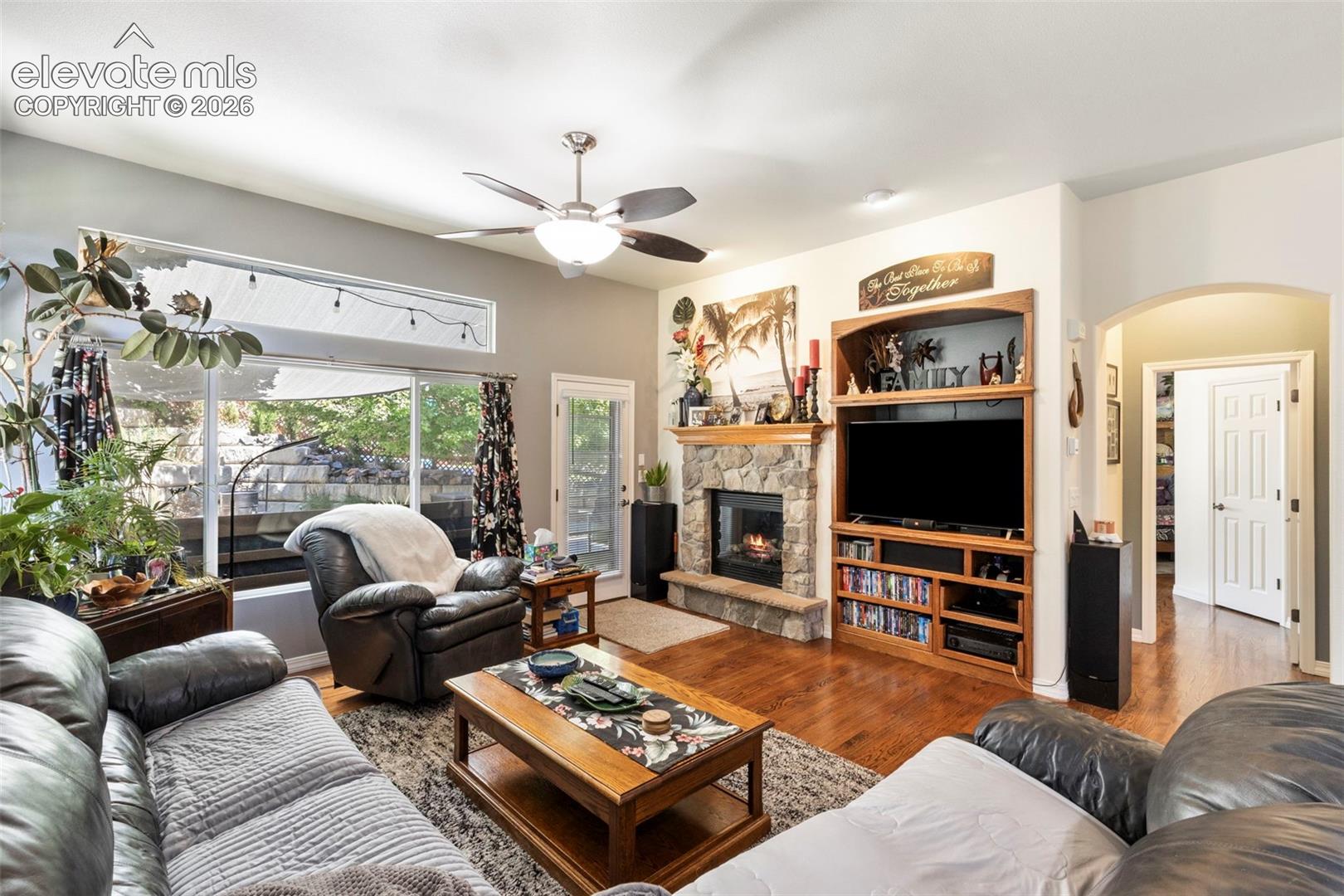 Wood-finish flooring, stone-front fireplace with hearth, built-in entertainment center, ceiling fan, and large windows