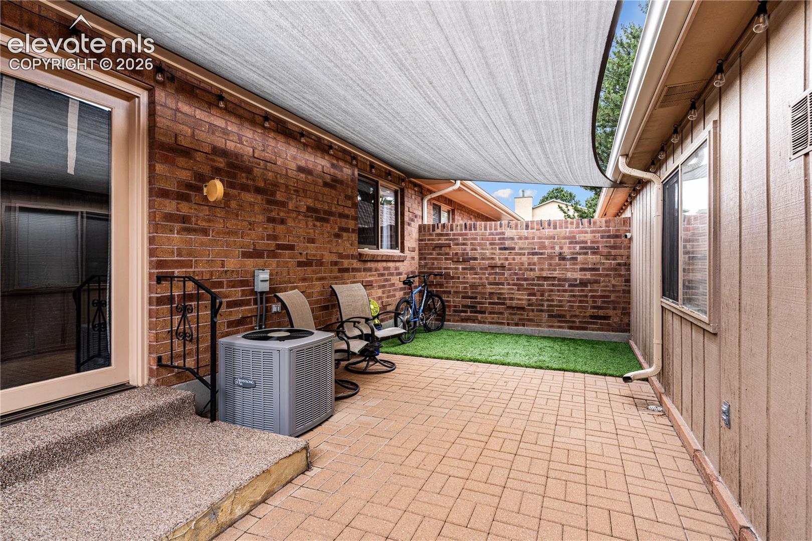 Outdoor patio featuring herringbone-patterned brick pavers, a fabric shade sail, an enclosed brick privacy wall, and a turf-grass area