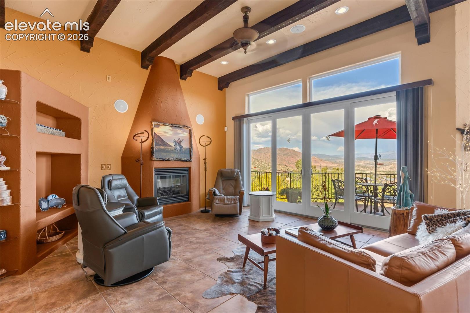Vaulted ceilings with exposed wood beams, kiva-style fireplace, large tile flooring, built-in recessed shelving, and expansive glass doors