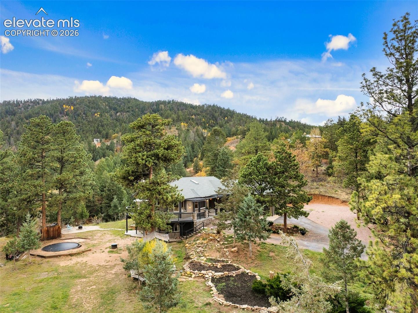 Multi-level residence featuring a wrap-around deck, metal roofing, stone-lined landscaping beds, and a gravel driveway, set against a mountainous backdrop