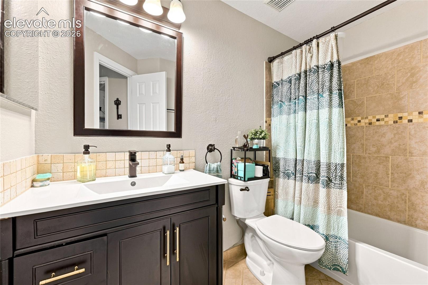 Dark-finish cabinetry featuring gold-toned hardware, white vanity countertop with an integrated rectangular basin, matte black faucet, framed vanity mirror with three-light fixture, and neutral tile tub surround with decorative mosaic accent inlay