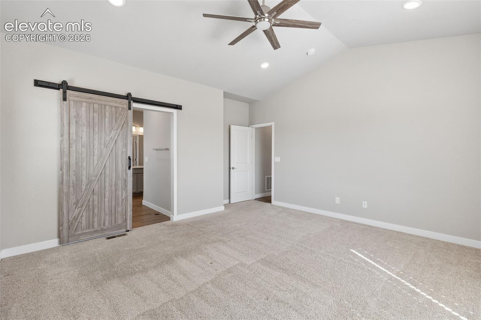Vaulted ceiling featuring a central multi-blade ceiling fan, neutral wall-to-wall carpeting, and a sliding barn-style door with matte black hardware