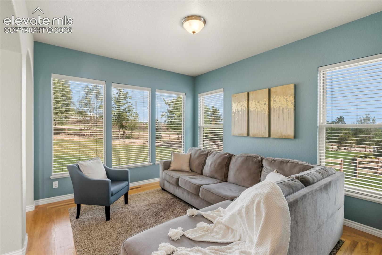 Bright living area featuring multiple large windows with horizontal blinds, wood-finish flooring, and a neutral wall color