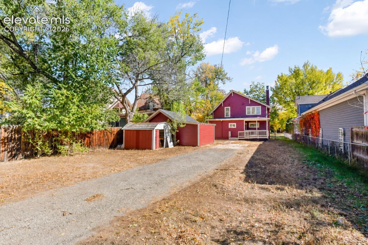Exterior view featuring a gravel driveway, detached storage structure, raised wooden deck with railings, and perimeter wood and chain-link fencing