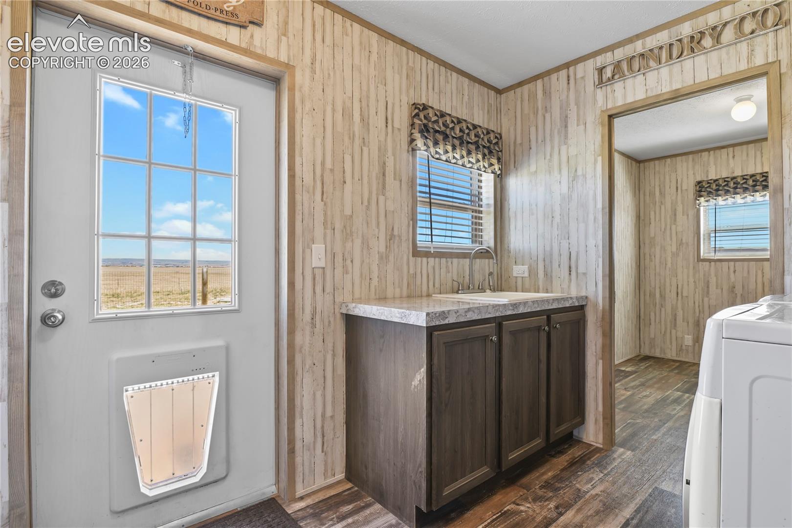 Utility space featuring wood-finish wall paneling, dark cabinetry with a stone-finish countertop, stainless steel drop-in sink, and exterior door with integrated window and pet portal
