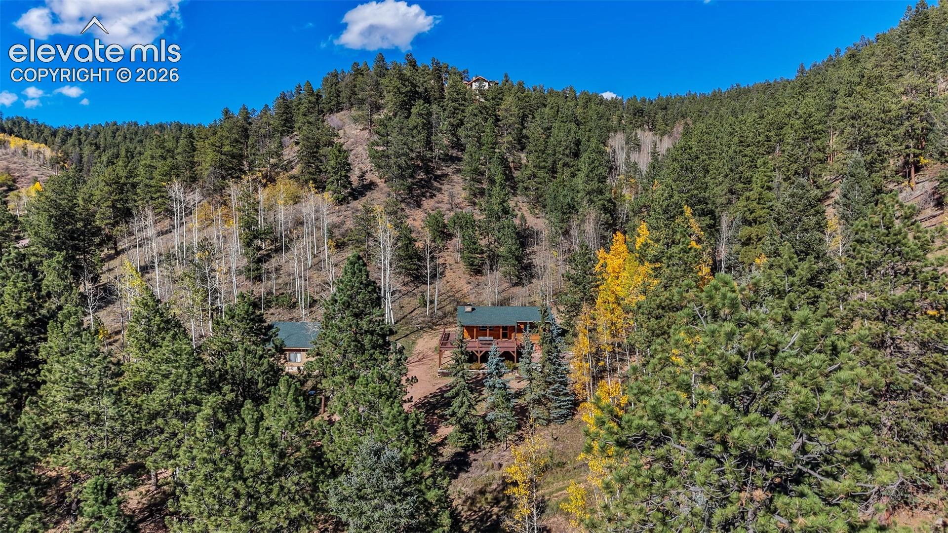 Log cabin structure situated on a sloped mountain terrain featuring a dark-toned gable roof, a wraparound wood deck, and dense surrounding evergreen and deciduous forest