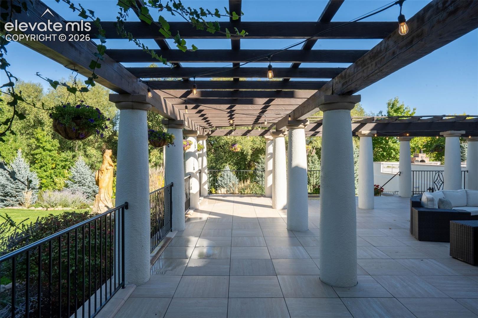 Covered terrace featuring a wood-beam pergola, stone-finish tile flooring, classic cylindrical columns, and a black wrought-iron railing